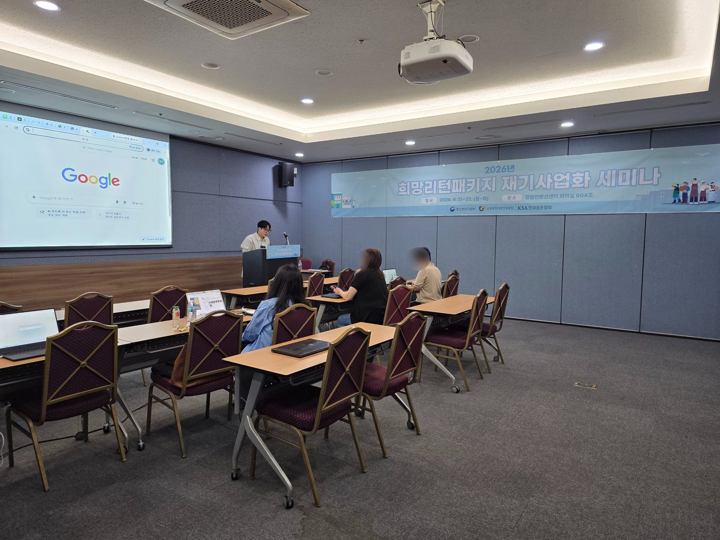 창원컨벤션센터 회의실에서 진행한 2026년 희망리턴패키지 재기사업화 세미나 현장. 오른쪽 벽에 세미나 배너가 걸려 있고 참가자들이 각자 노트북을 펴고 앉아 있다